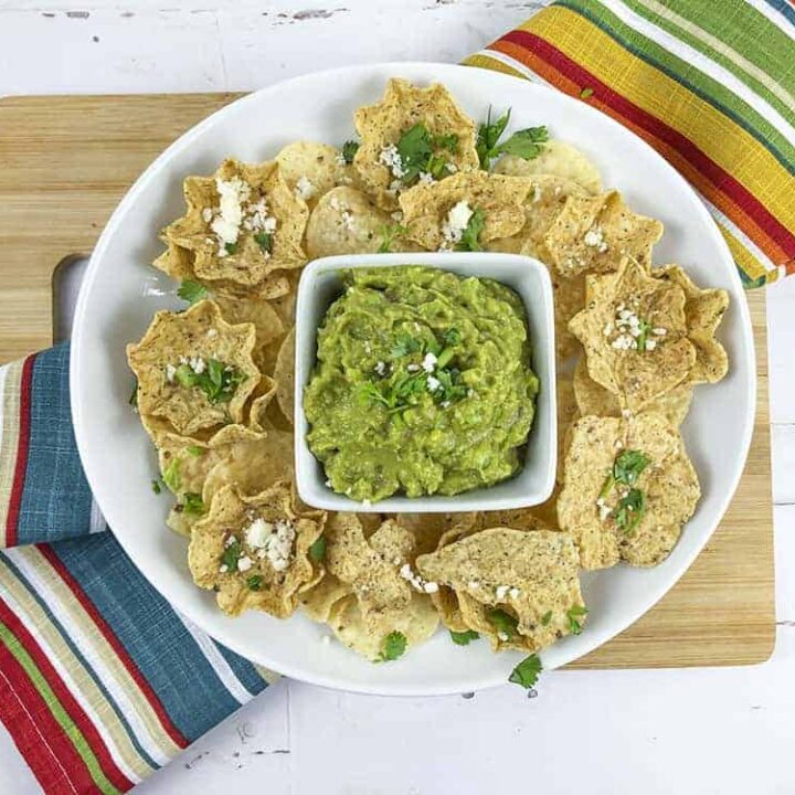 An overhead image of guacamole sitting in a square dish with tortillas surrounding it. There is a colorful table cloth laying underneath the white plate.