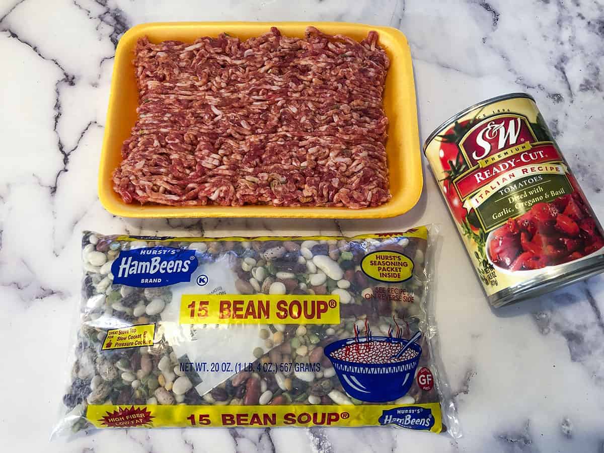 Raw sausage, a can of diced tomatoes and a package of 15 bean soup mix on a marble table.
