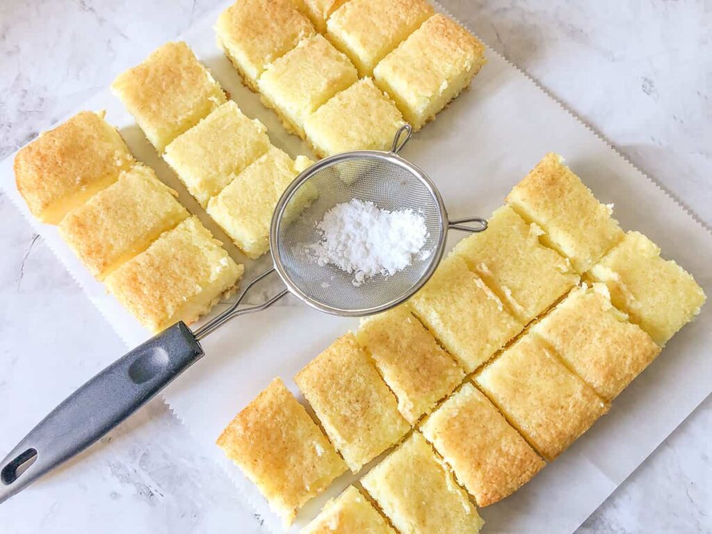 Lemon bars that have been baked and cut, with a sieve full of icing sugar for dusting in the middle.
