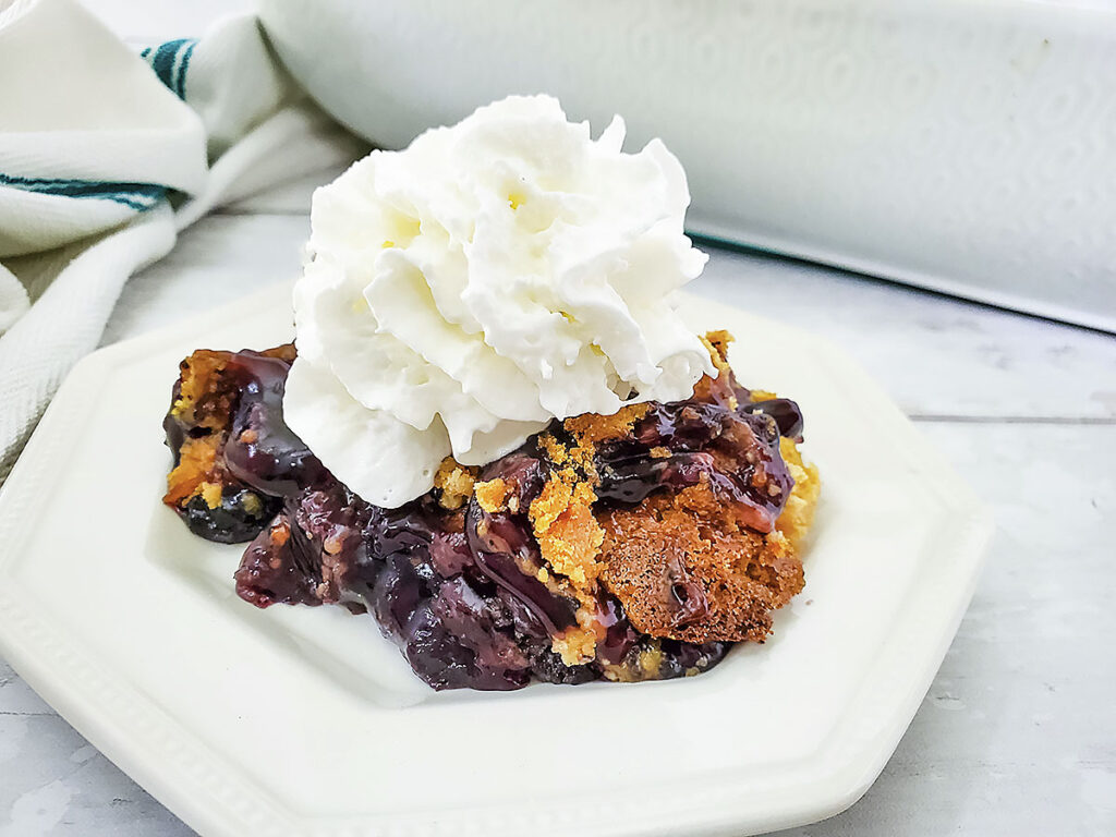 Blueberry dump cake on a white plate with whipped cream over top the cake.