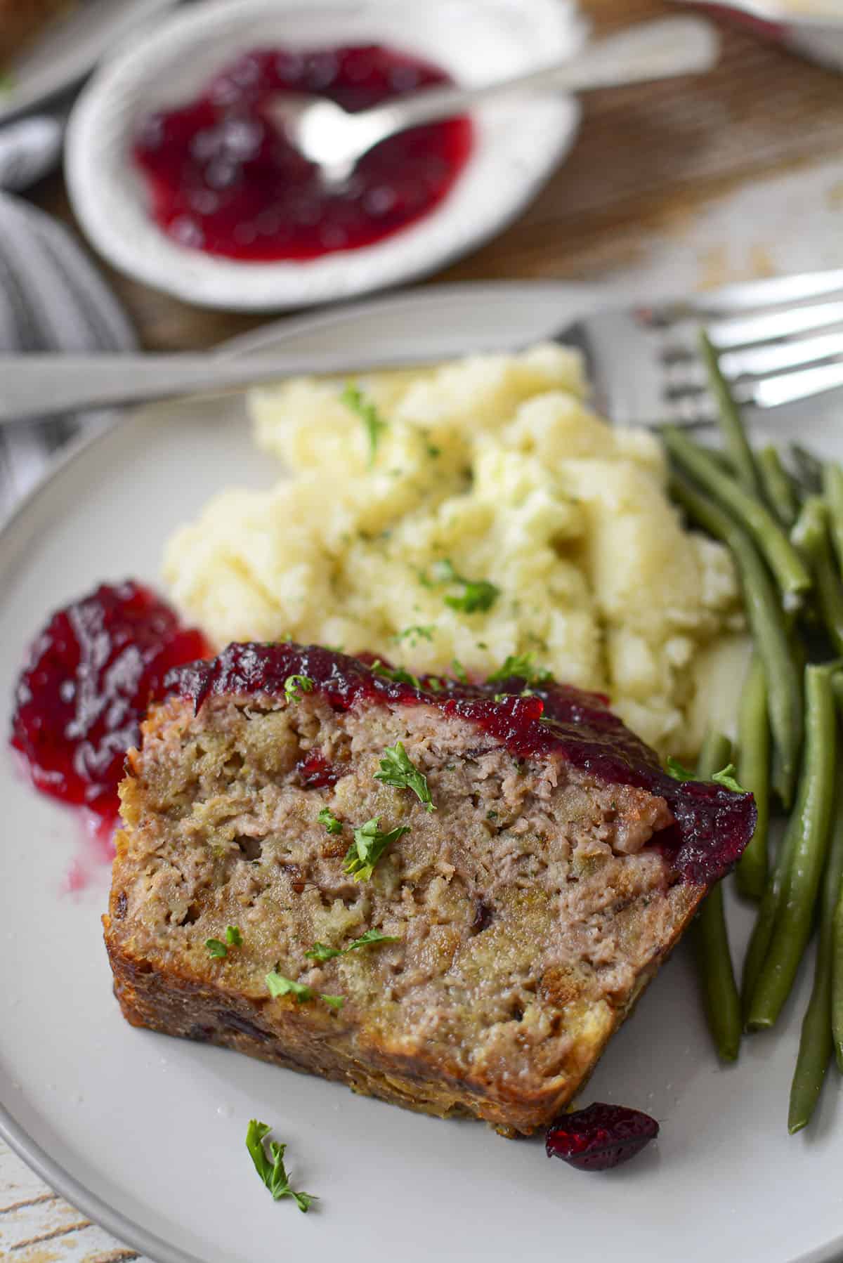 One piece of meatloaf resting on mashed potatoes and green beans are on the right. A dollop of sauce is on the left.