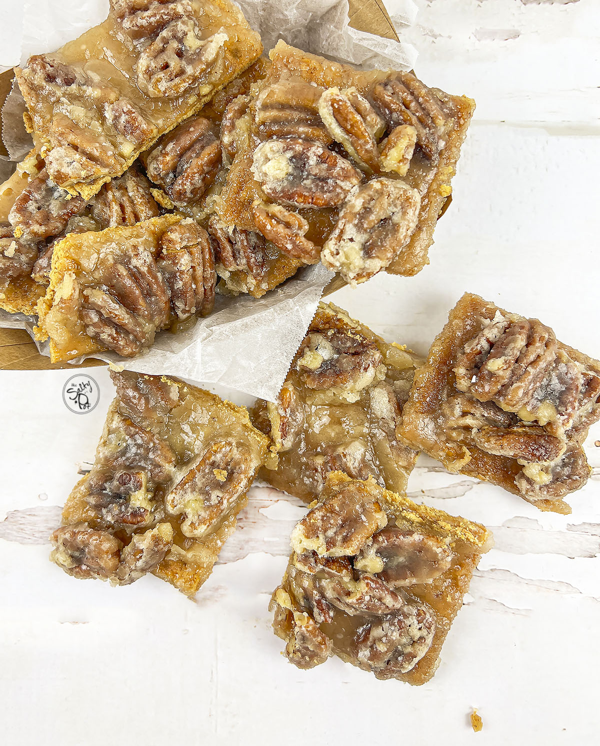 Pecan pie bark in a cardboard container on the upper right with four pieces on the table in front of it.