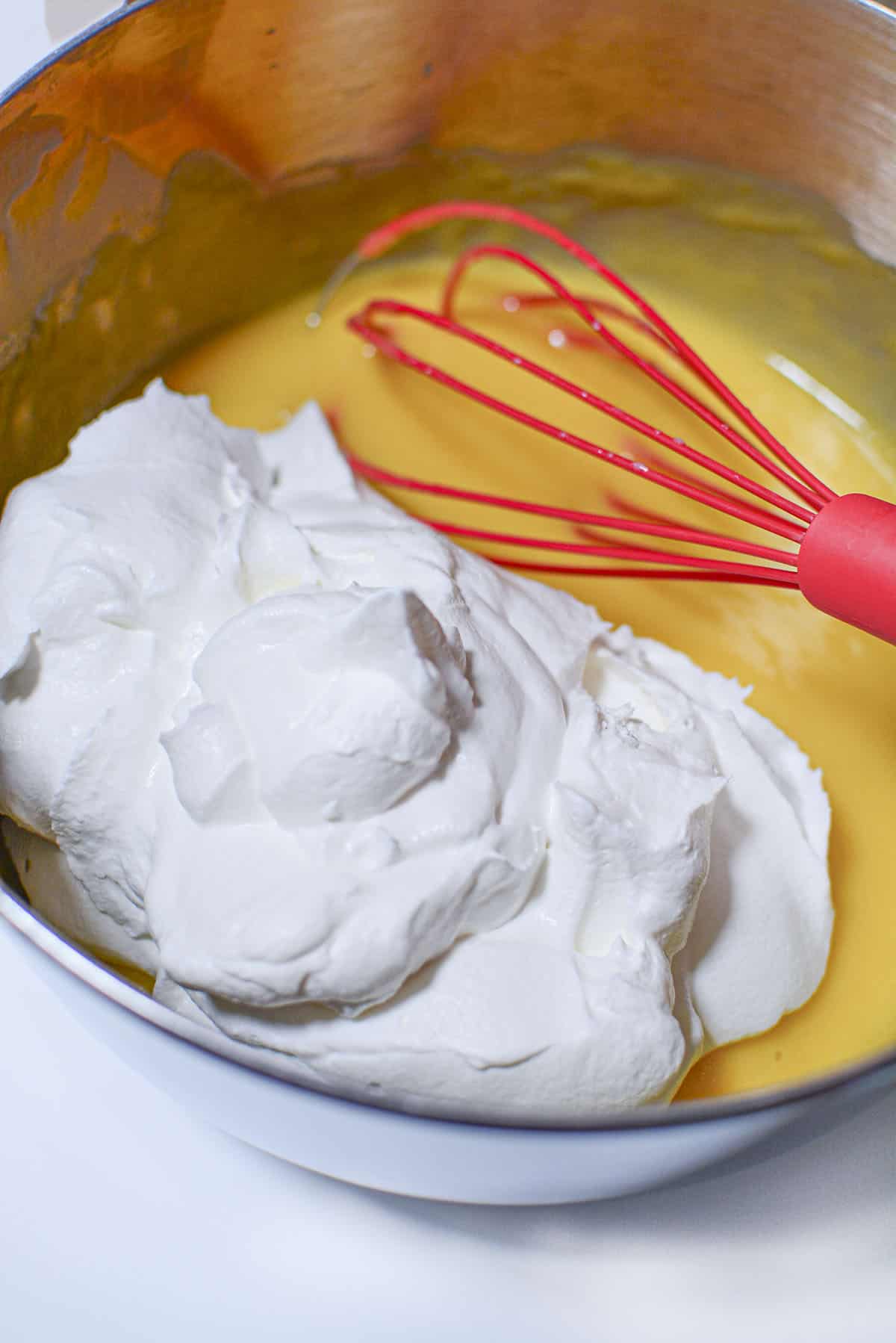Whipped cream is about to be folded into the eggnog pudding. There is a large red whisk next to the whipped cream.