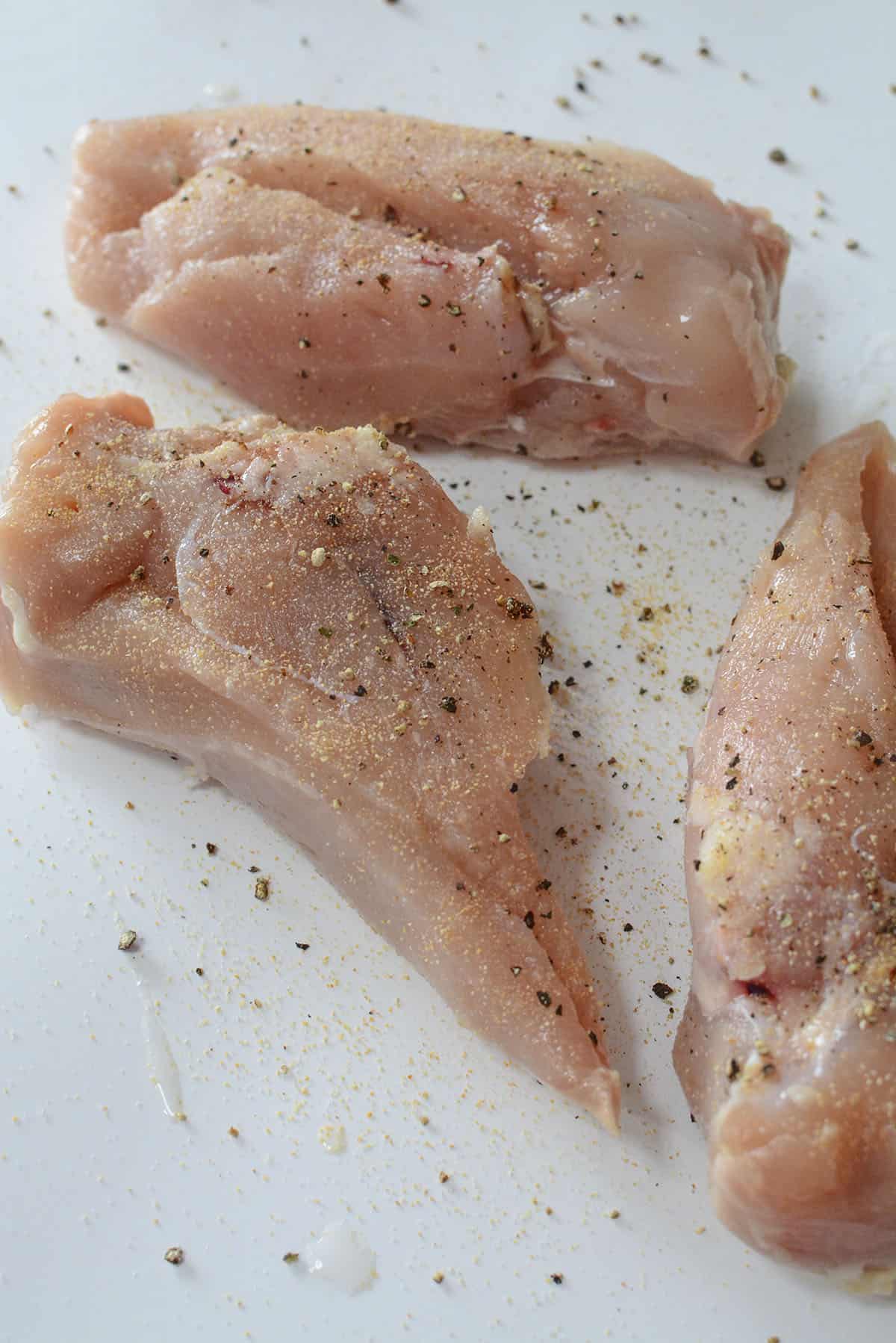 3 seasoned chicken breasts on a white plastic cutting board.