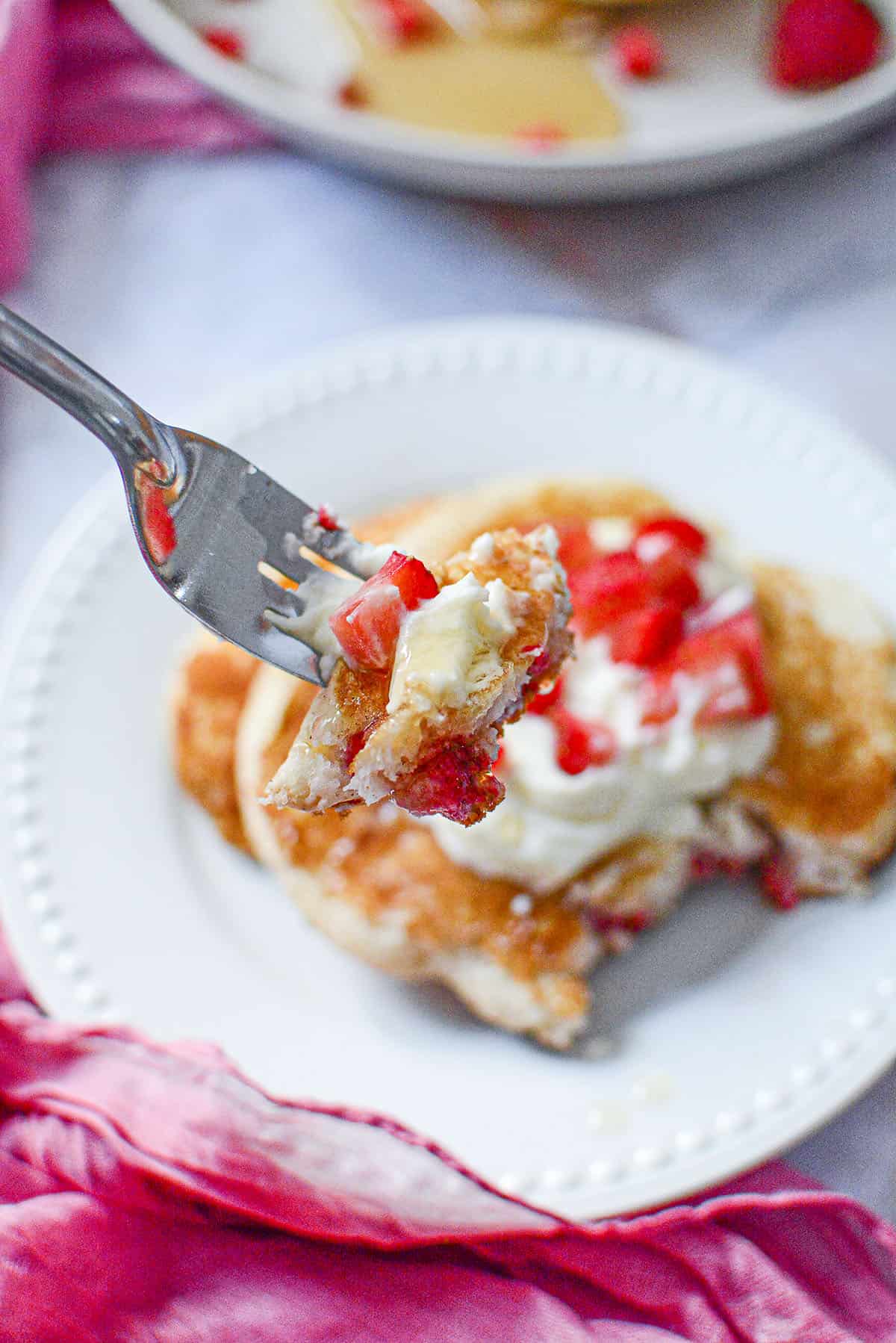 A fork with a mouthful of this cooked strawberry pancake recipe. Fresh strawberries along with whipped cream are on the fork as well. 