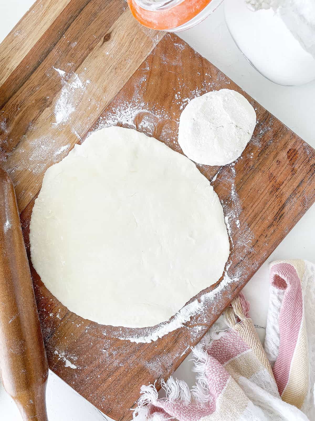 The dough ball is rolled out thin. The rolling pin is on the left side with a towel at the bottom of the cutting board.