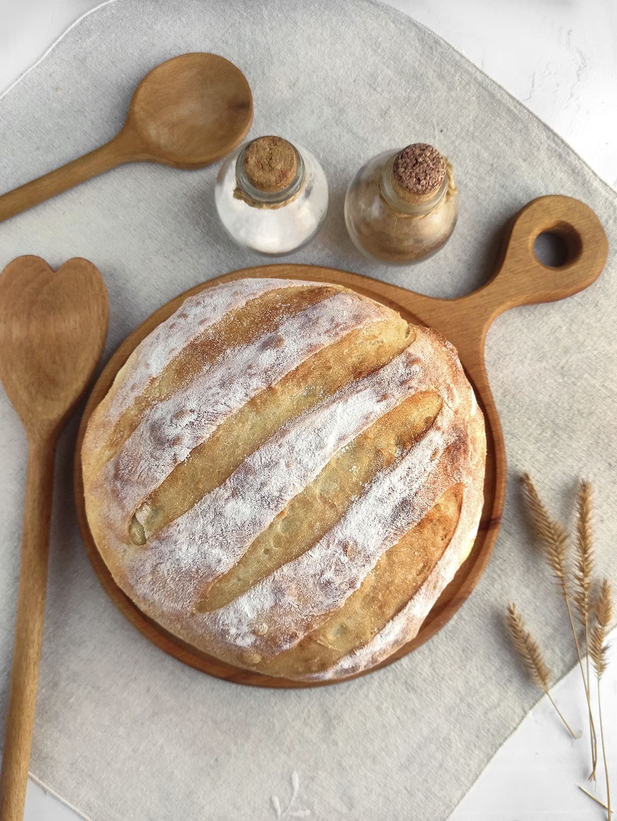 A freshly baked loaf of round bread on a grey napkin with spoons and other garnishes around it. 