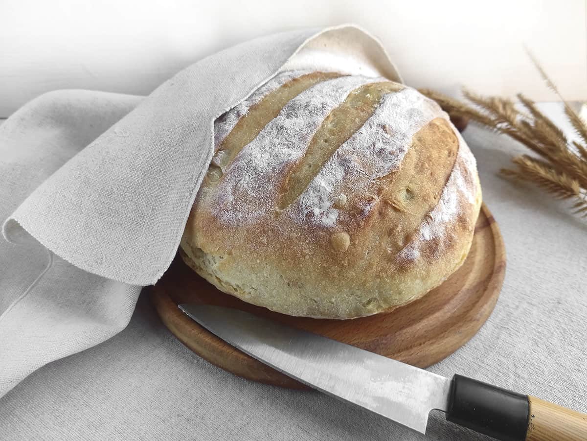 The round artisan type bread loaf, unsliced, with a cooling linen towel over half of it. A knife sits at the front, ready to cut into slices.