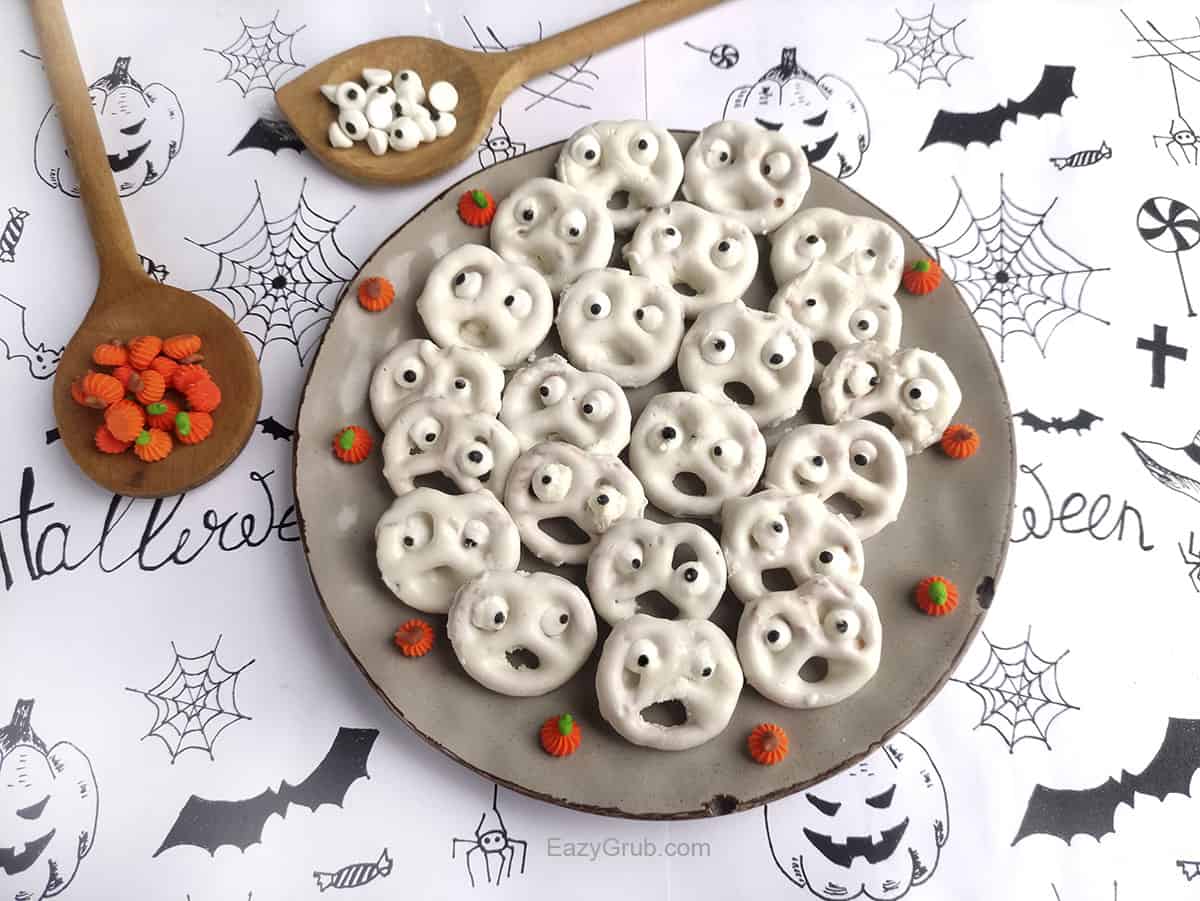 22 pretzel ghosts on a plate with a halloween background. Two wooden spoons hold candy on the upper left of the plate.