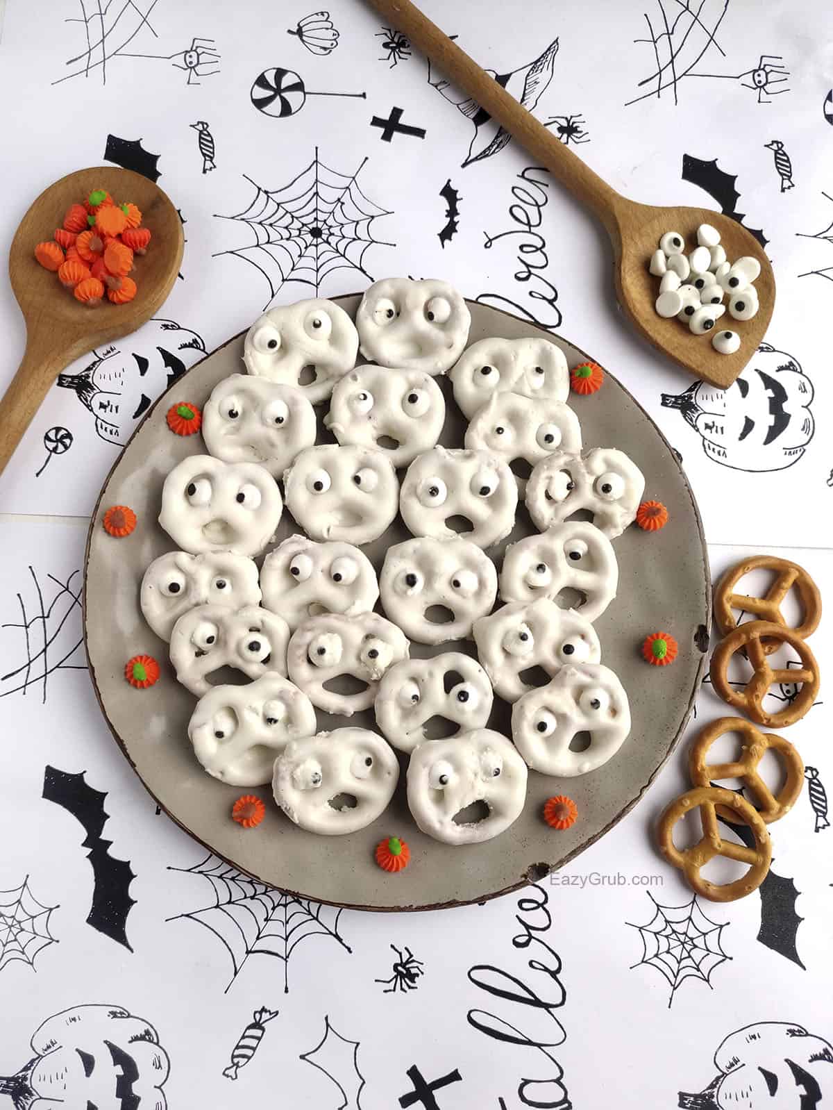 Pretzel ghosts sit on a plate with pretzels on the bottom right, and wooden spoons filled with candy pumpkins and candy eyes above the plate.