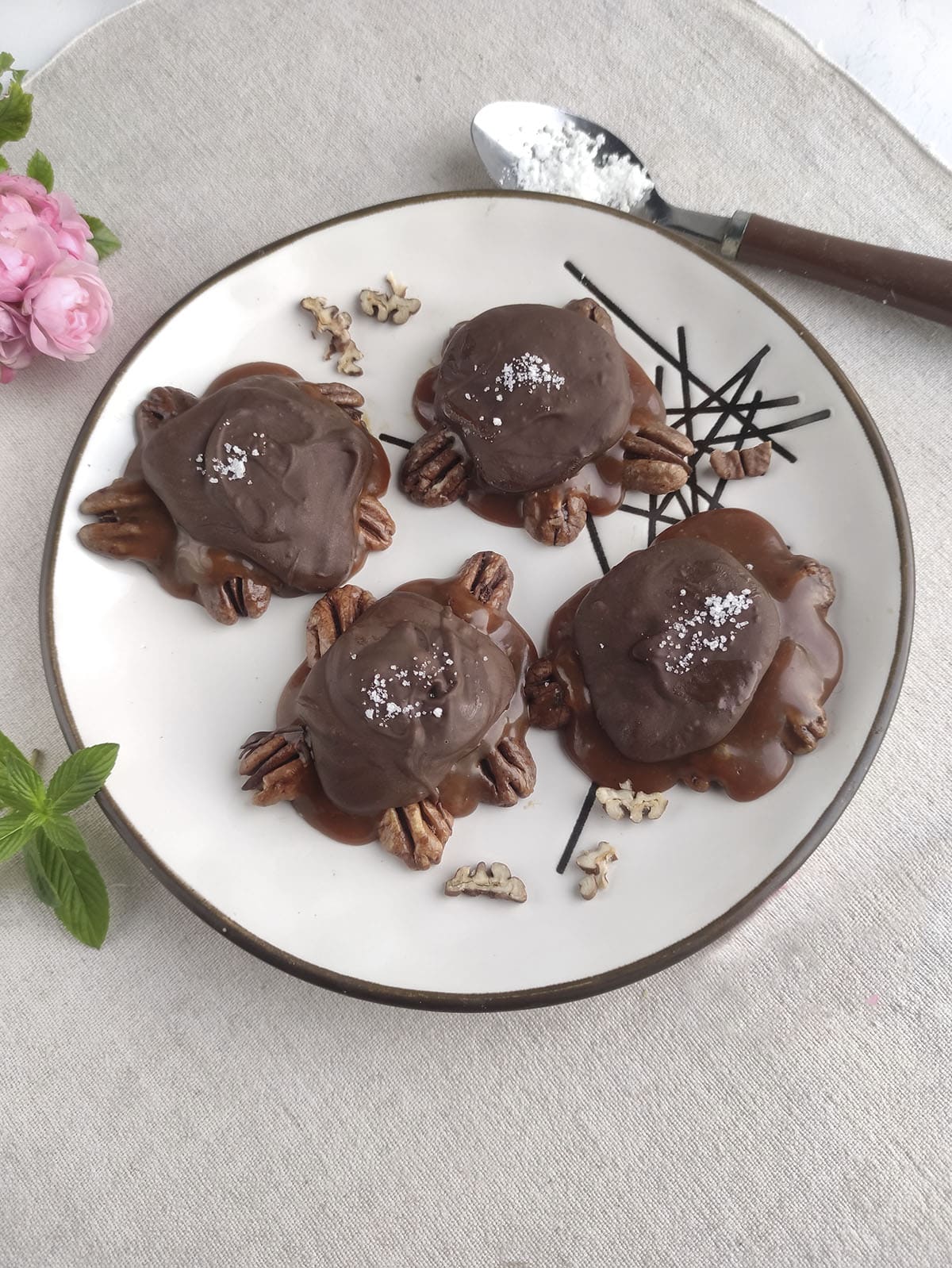 4 chocolate pecan turtles on a plate with a small spoon filled with salt on the upper right.