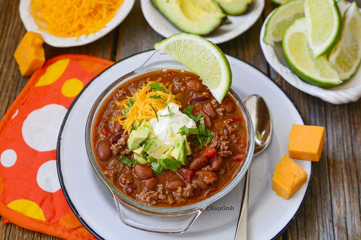Chili with cheddar cheese, sliced avocado and lime wedges in bowls surrounding it. There is an orange and polka dot napkin under the plate.