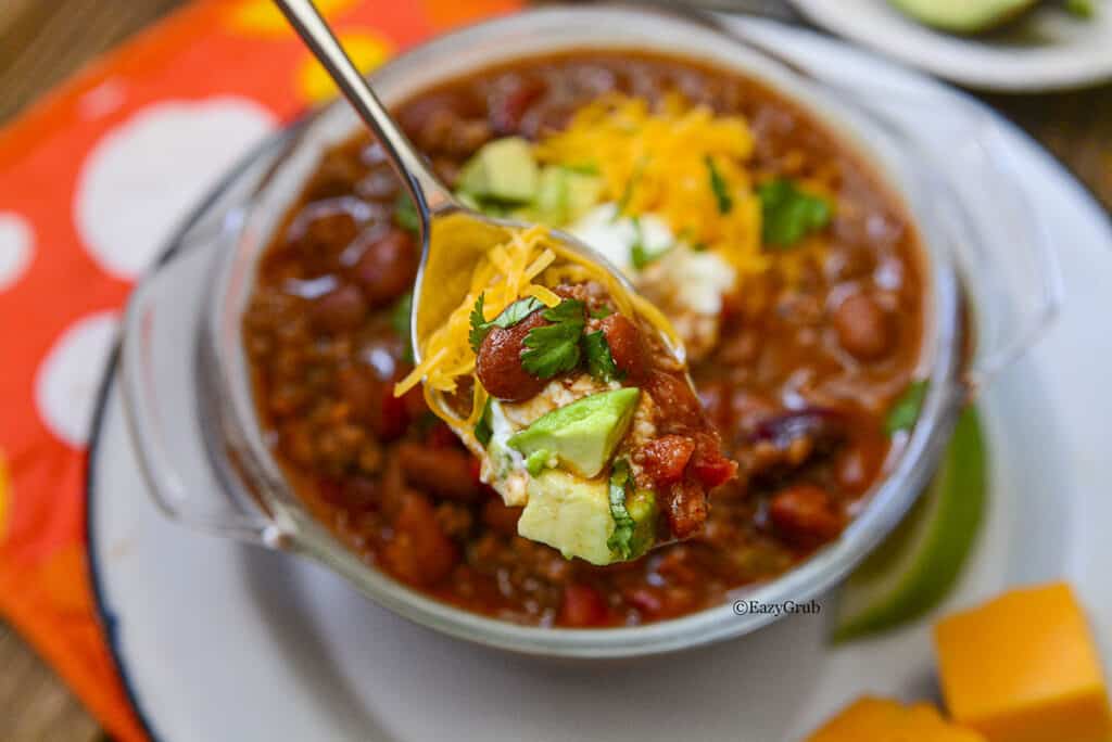 A spoon with chili and toppings on it, hovering over a bowl. 