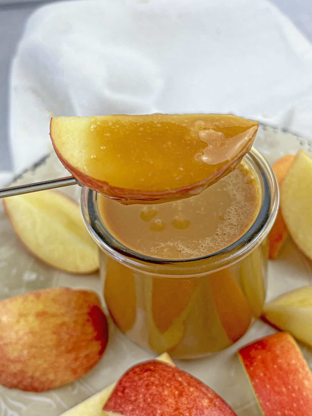 A slice of apple that has been dipped in the caramel, hovering above the jar of caramel.