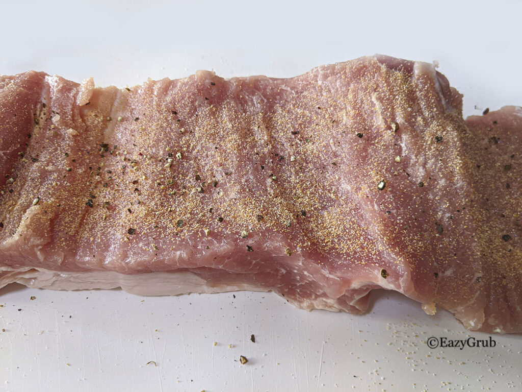 A butterflied pork loin roast, seasoned, on a white plastic cutting board.