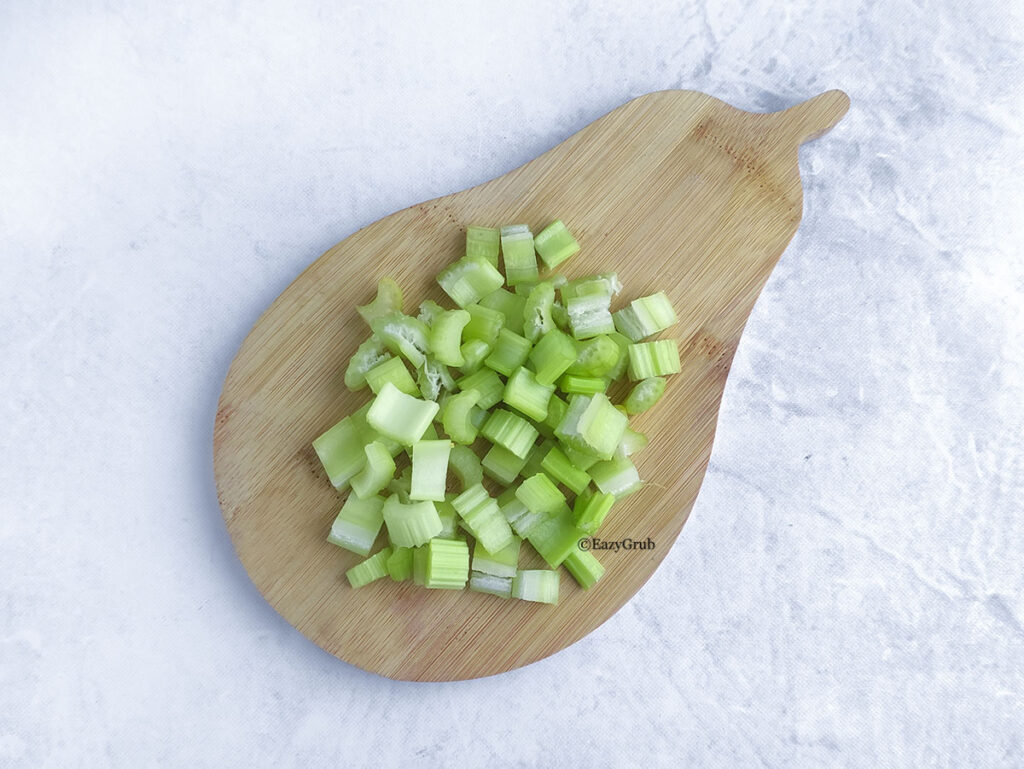 A neat pile of finely diced green celery stalks centered on a unique, pear-shaped wooden cutting board, resting on a light-colored surface.