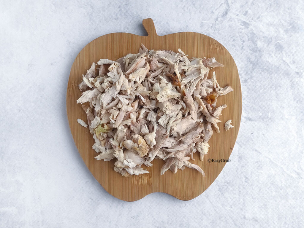 A large mound of shredded roasted chicken meat is piled high on an apple-shaped wooden cutting board, resting on a white marble-patterned surface.