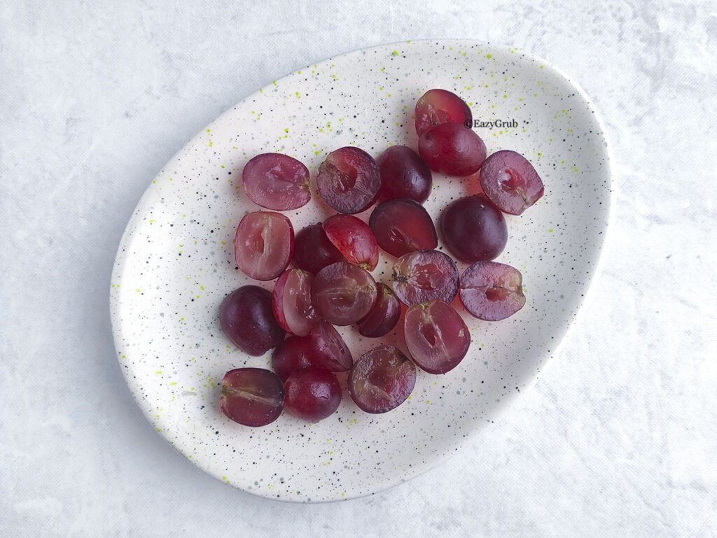 Approximately twenty-five halved red grapes are spread out on a speckled, oval-shaped white ceramic plate, resting on a white patterned background.