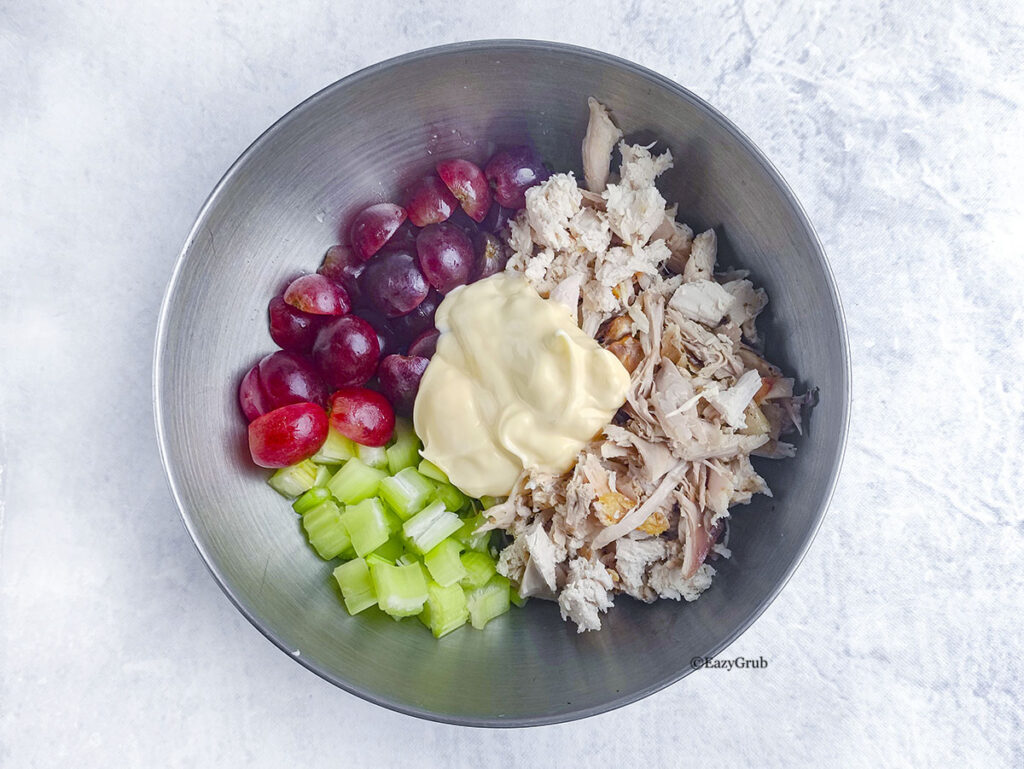 A close-up looking down into a stainless steel mixing bowl, where shredded rotisserie chicken, halved red grapes, diced green celery, and a large dollop of mayonnaise are ready to be mixed together.