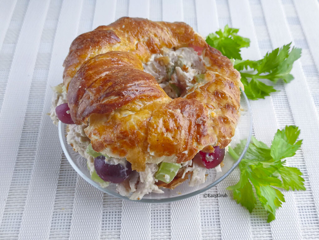 A side profile view of the completed chicken salad sandwich inside a croissant-bagel, showing the generous layer of filling between the top and bottom halves of the flaky pastry.