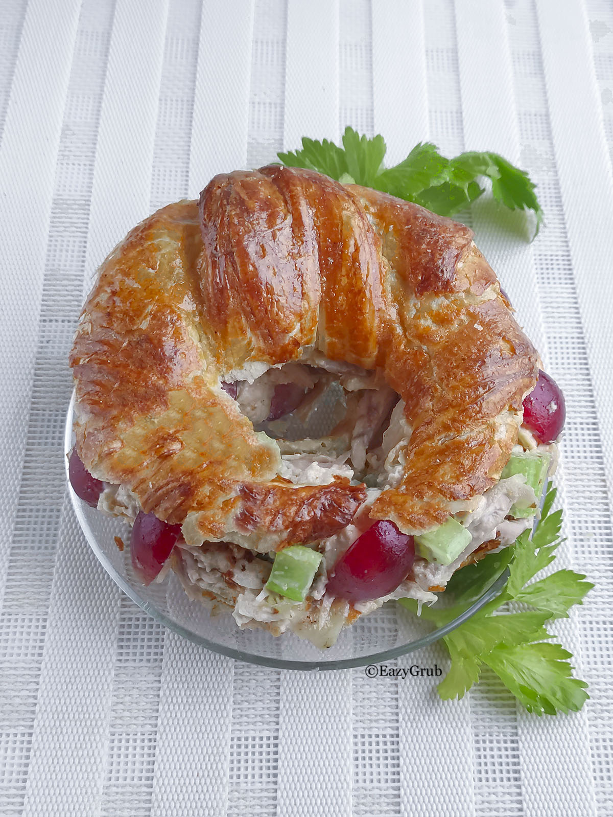 A detailed close-up of the finished chicken salad served inside a flaky, golden-brown croissant-bagel, resting on a small glass plate with fresh celery leaves as garnish.