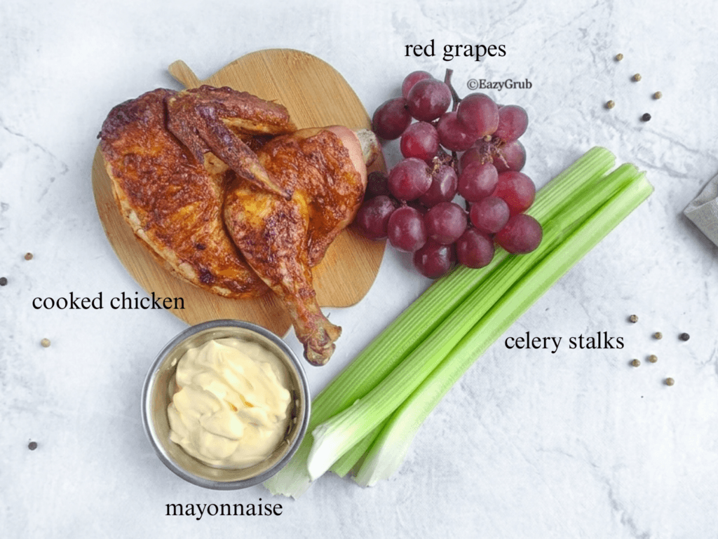 An overhead view of the 4 ingredients used in the recipe, laid out on a light grey background: a cooked whole roasted chicken on an apple-shaped cutting board, a large bunch of red grapes, several crisp celery stalks, and a small metal bowl filled with creamy mayonnaise.