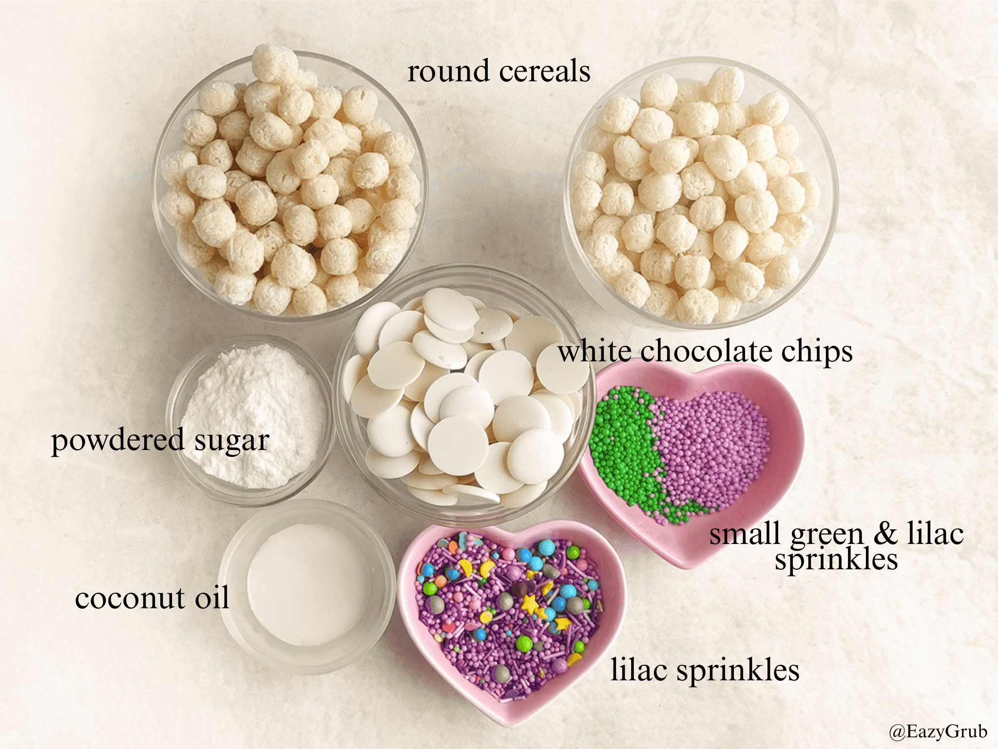 Ingredient image: white chocolate chips, round cereal, coconut oil, lilac sprinkles, green and lilac sprinkle balls, and powdered sugar each in separate bowls on a countertop