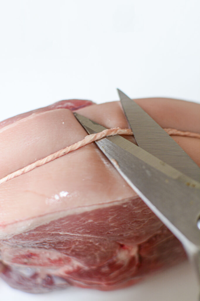 A close-up shot of a pair of metal scissors cutting through the elastic netting of a raw pork shoulder roast to prepare it for seasoning.
