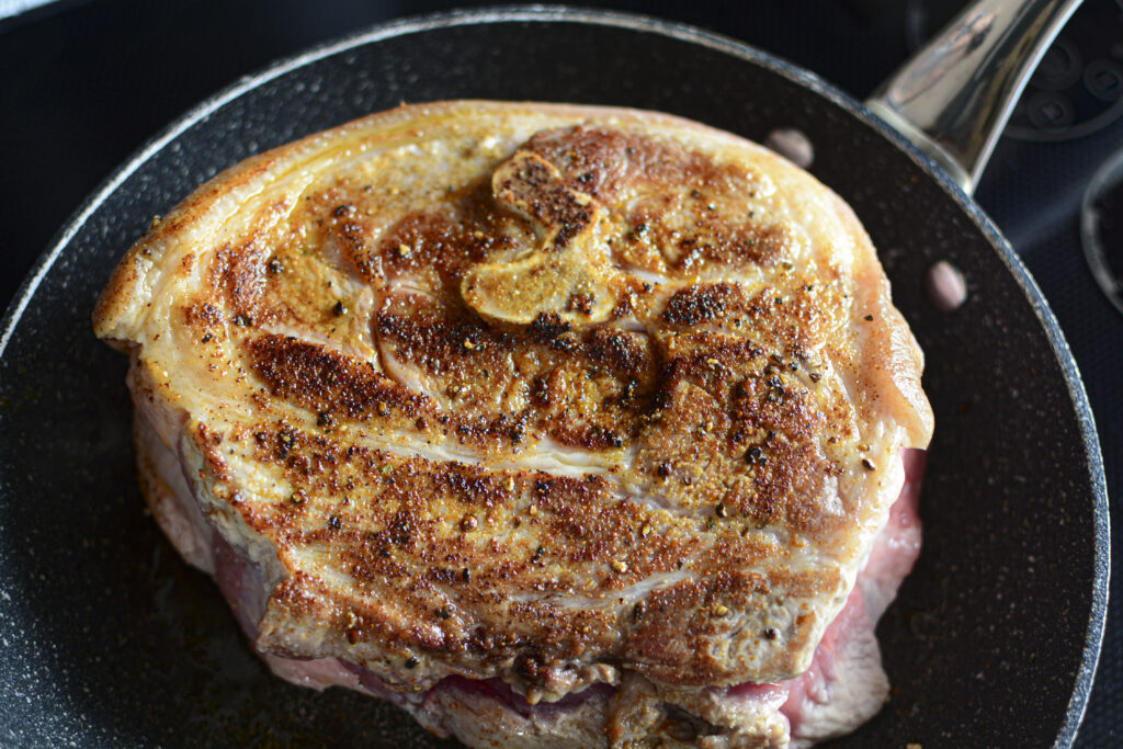 The seasoned pork roast being seared in a black non-stick skillet. The top surface of the meat is golden-brown and caramelized from the heat.