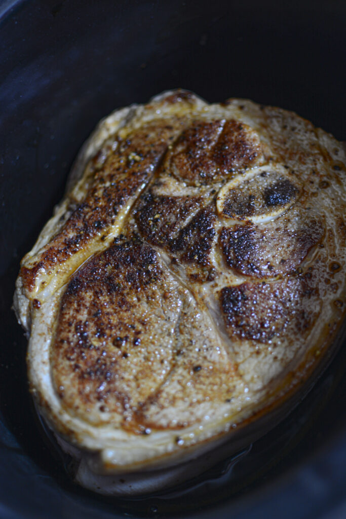 The seared pork roast placed inside the dark ceramic insert of a slow cooker, showing the browned crust and rendered juices at the bottom.