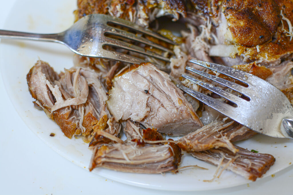 A close-up of the cooked pork on a white plate being shredded with two silver forks, revealing the tender, juicy interior of the meat.