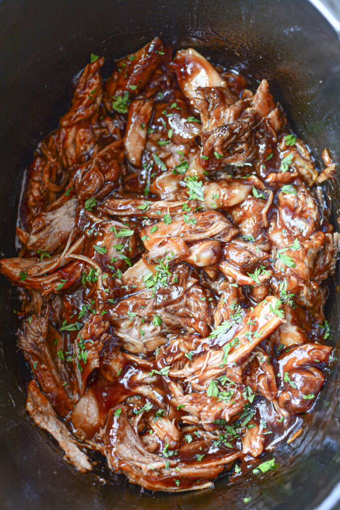 A vertical close-up shot of the shredded BBQ pulled pork inside the slow cooker, showing the thick sauce clinging to the meat fibers and a garnish of chopped parsley.