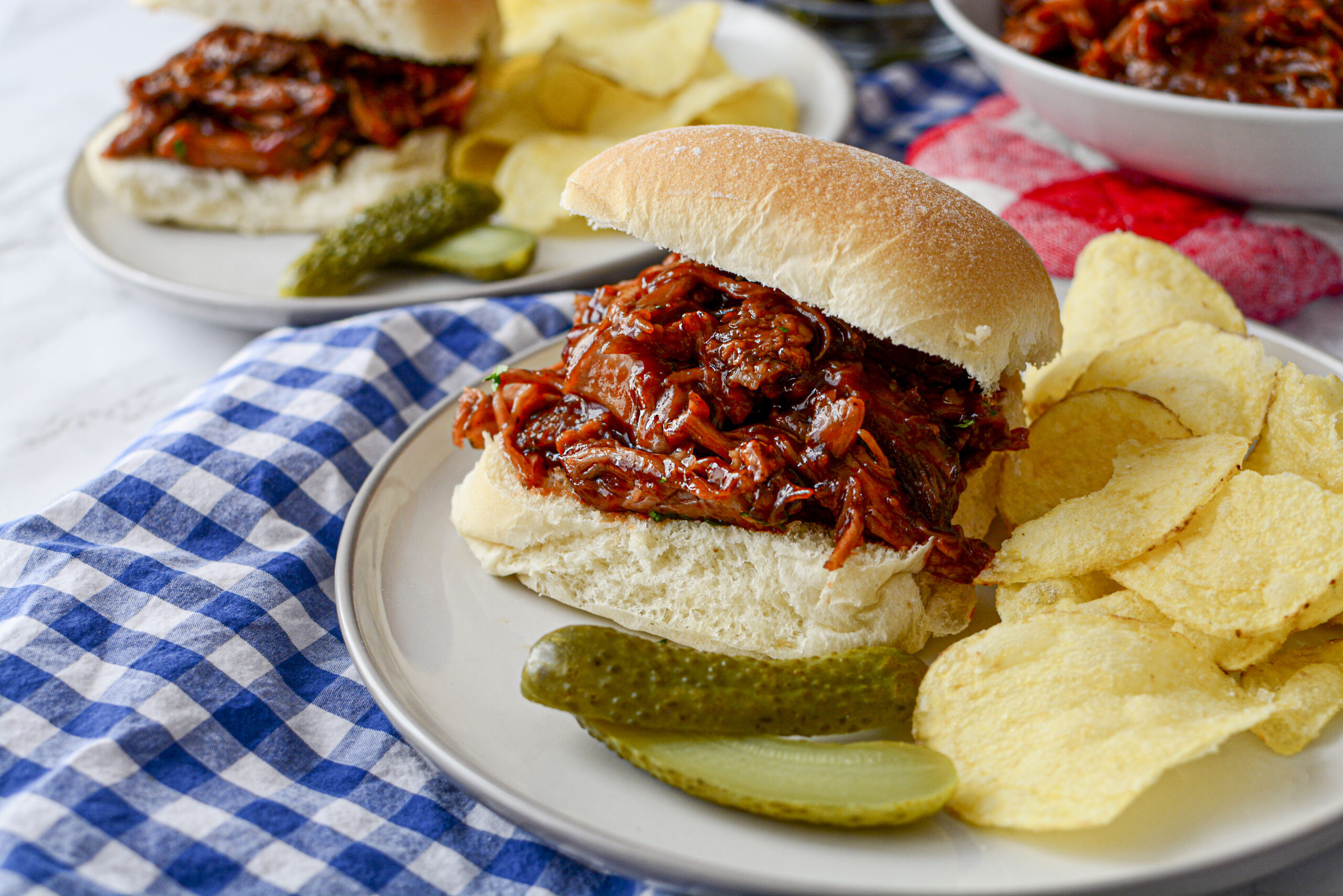 A finished BBQ pulled pork sandwich served on a white bun, placed on a light-colored plate with a side of potato chips and two pickle spears. The plate sits on a blue and white checkered napkin.