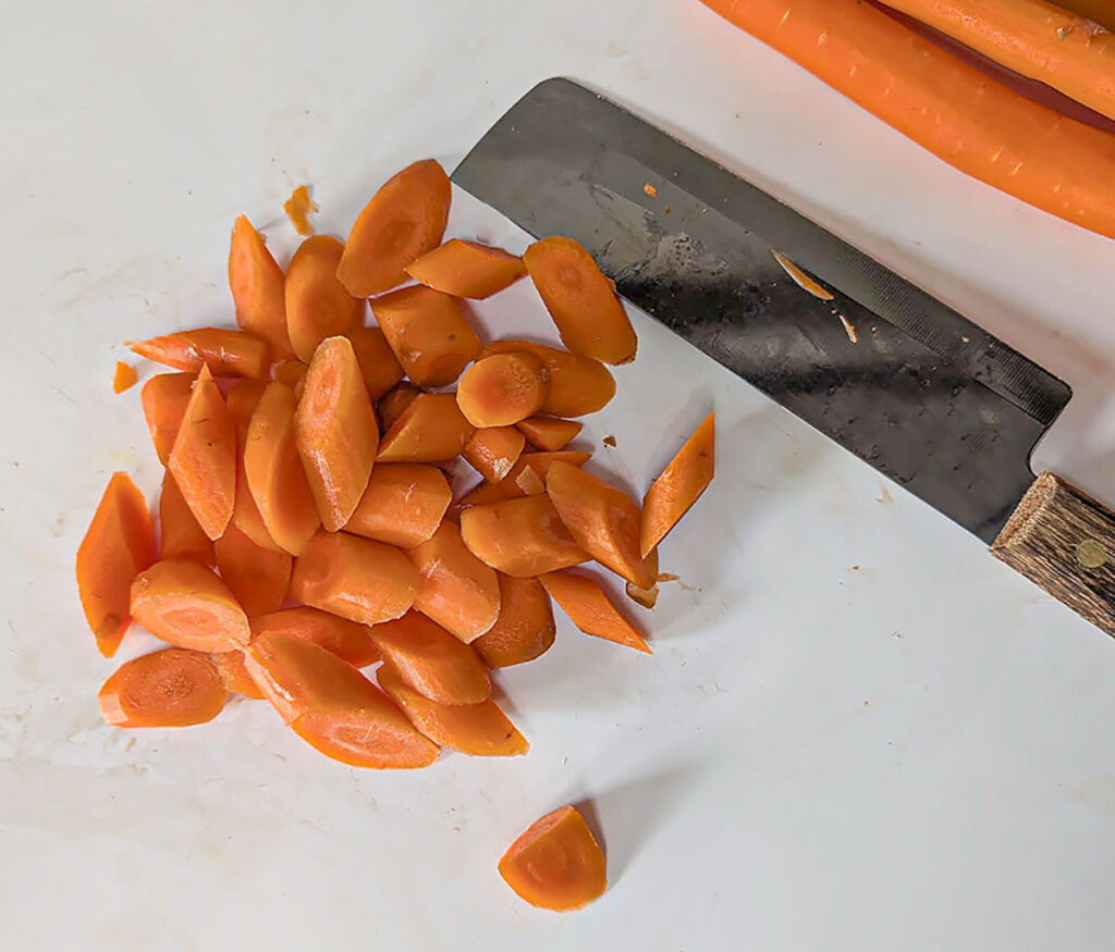 A close-up of bright orange carrots sliced into thick diagonal coins on a white cutting board next to a large kitchen knife.