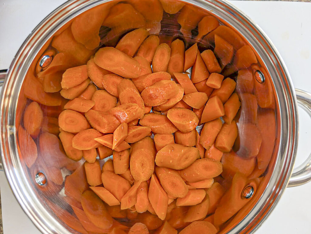 A top-down view looking into a stainless steel pot filled with a large batch of sliced raw orange carrots ready to be cooked.