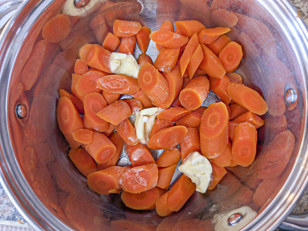 Sliced carrots in a stainless steel pot with three generous pats of yellow butter placed on top, beginning to melt.