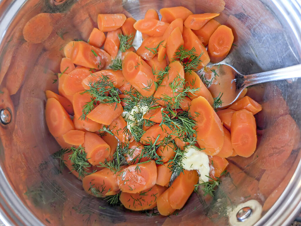 Cooked carrot slices in a pot being tossed with melted butter and a heavy garnish of freshly chopped green dill.