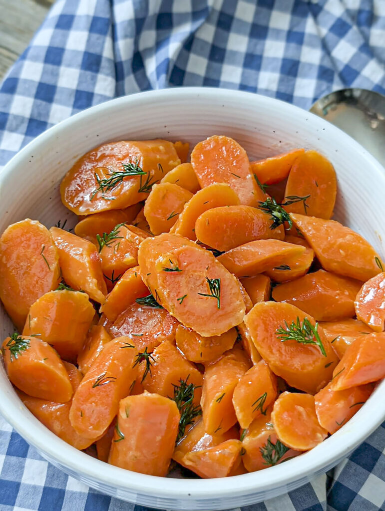 A vertical close-up shot of a white bowl filled with buttery dilled carrots, showing the glossy texture and fresh herbs against a blue and white checkered napkin.