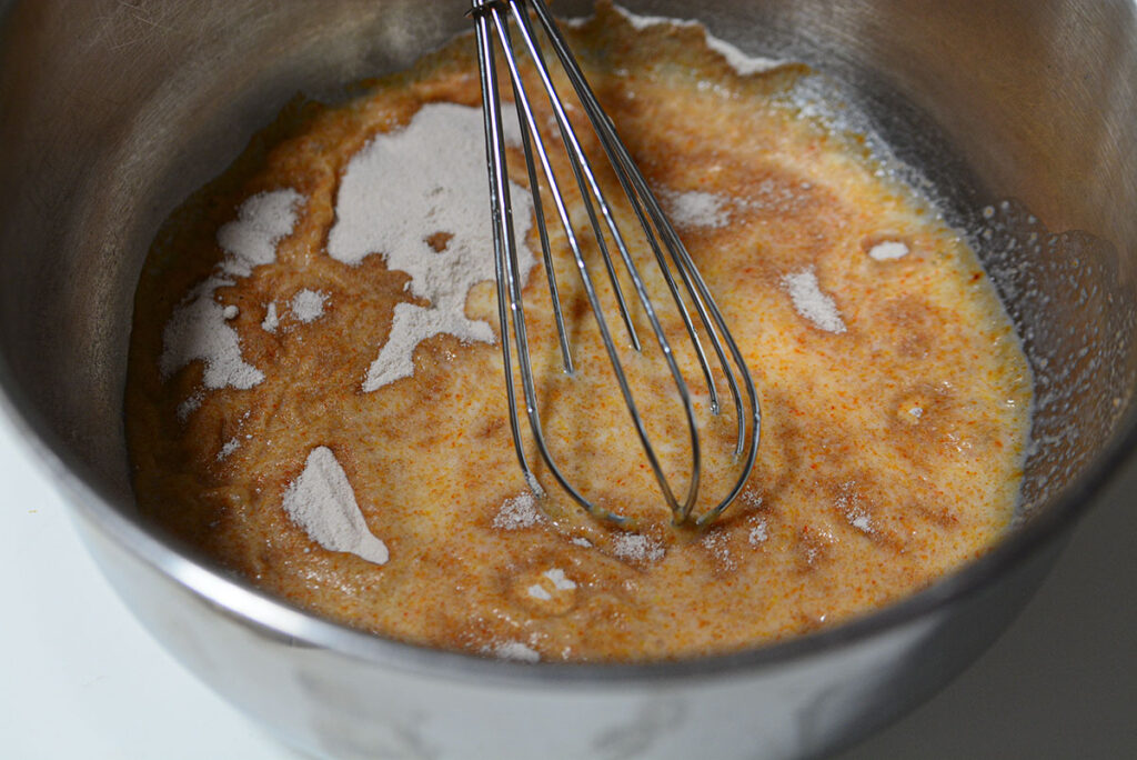 A high-angle, close-up shot of a stainless steel mixing bowl. Inside is a wet, orange-tinted base mixture being whisked. Several small piles of light brown powdered pudding mix are scattered across the surface.
