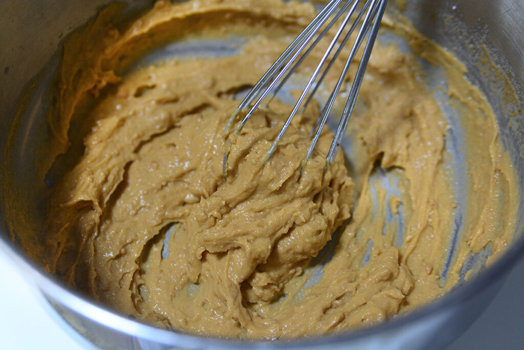 A close-up of the mixing bowl where the ingredients have been combined into a thick, smooth, caramel-colored paste. A wire whisk is resting in the center of the mixture.