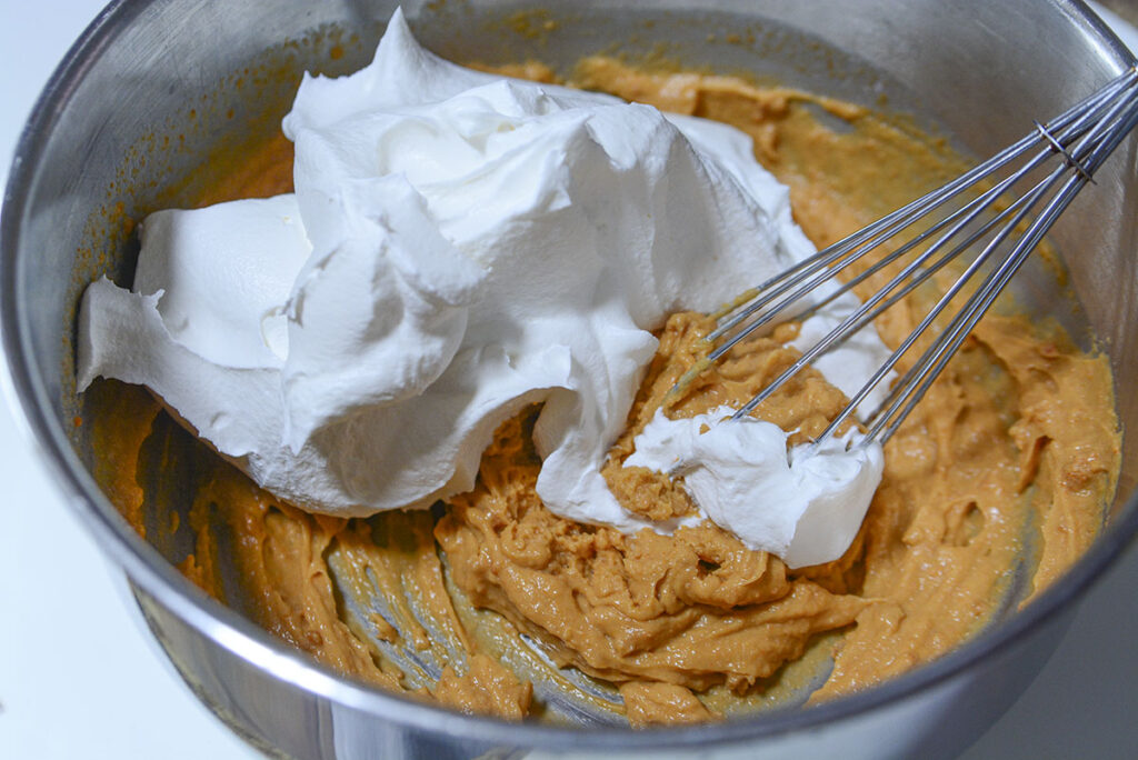 A large, fluffy mound of white whipped topping is being added to the thick butterscotch base in the metal bowl.