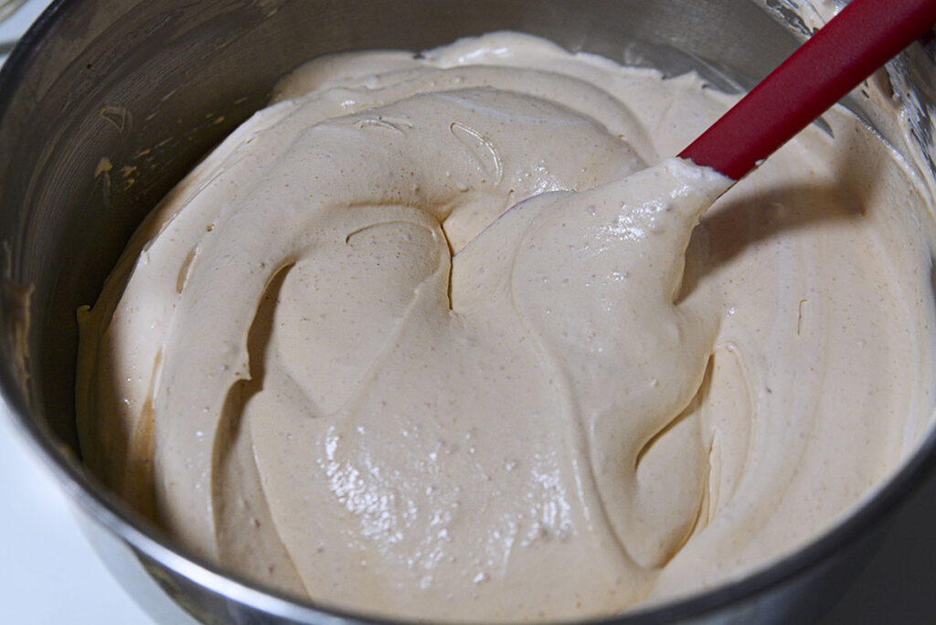The final filling mixture, now a light beige and airy mousse-like consistency, is being smoothed over with a red rubber spatula in the mixing bowl.
