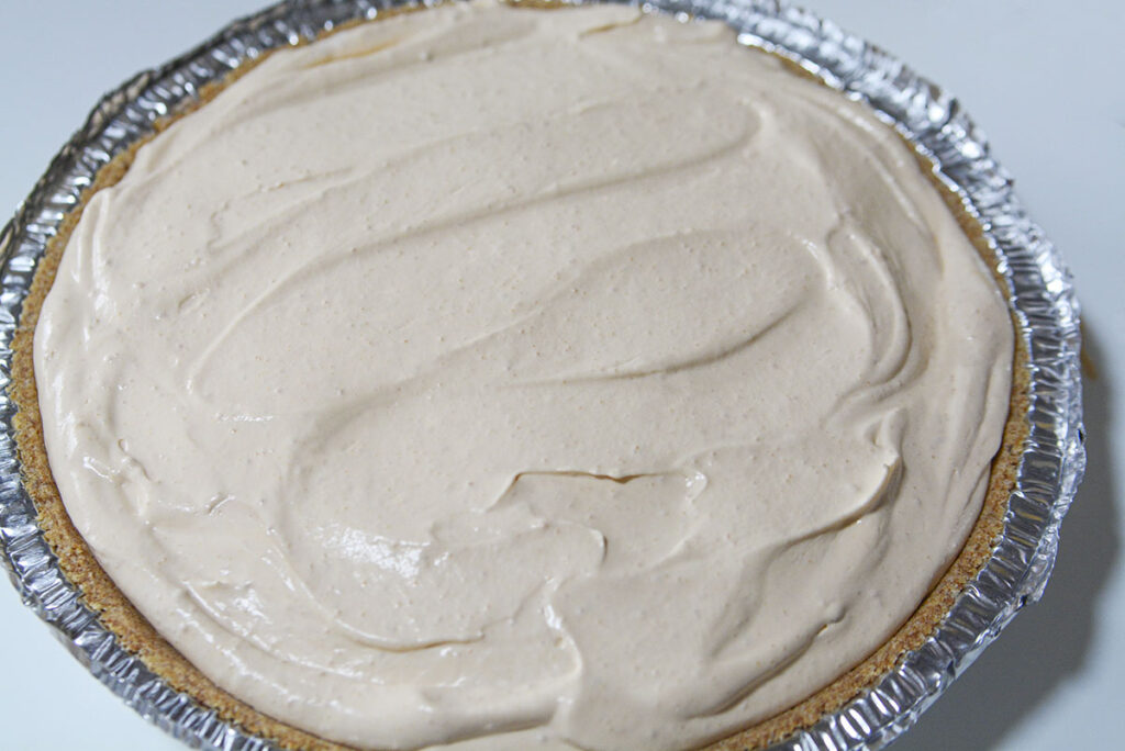 The light brown butterscotch filling has been poured into a graham cracker crust and leveled off. The pie sits in a disposable aluminum tin.