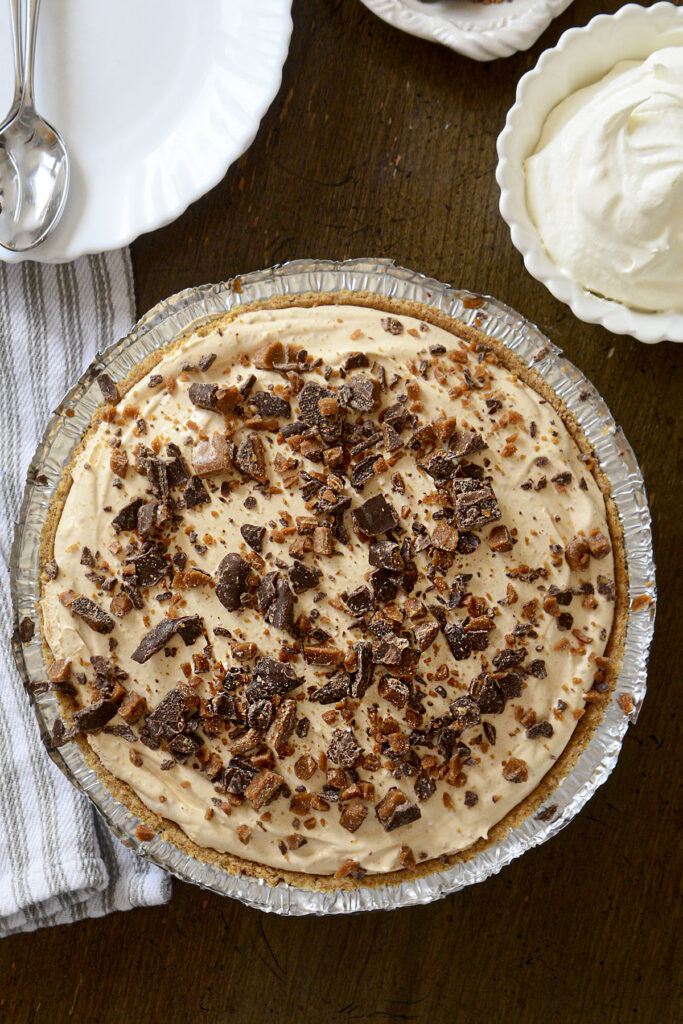 An overhead shot of a completed Butterscotch Pie in a graham cracker crust. The light tan, creamy filling is topped with a generous layer of crushed toffee bar pieces, showing bits of chocolate and golden toffee. The pie sits on a dark wooden table next to a white bowl of fluffy whipped cream, a clean white plate with a silver spoon, and a striped linen napkin.