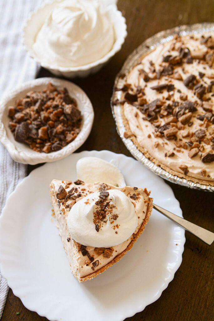 A bright, appetizing shot of a single slice of pie served on a white plate. The slice is topped with a swirl of whipped cream and more toffee bits. The whole pie and a small bowl of crushed candy are in the background.