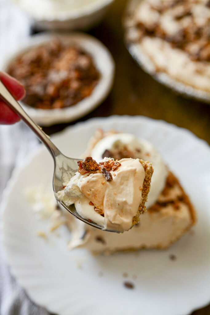 A close-up "action shot" of a silver spoon lifting a bite of the pie. The bite shows the light mousse texture, the crunchy toffee topping, and the graham cracker crust.