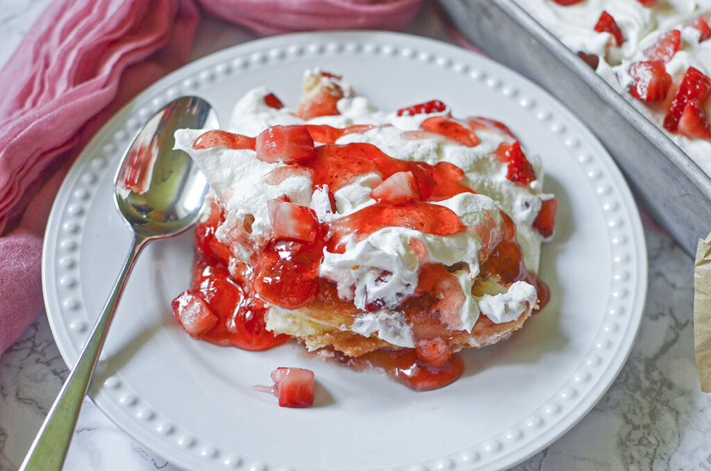 A close-up side view of a single square slice of the Twinkie strawberry cake served on a white plate with a silver fork, cleanly showcasing the distinct layers of golden cake, red strawberry glaze, and fluffy white whipped cream.