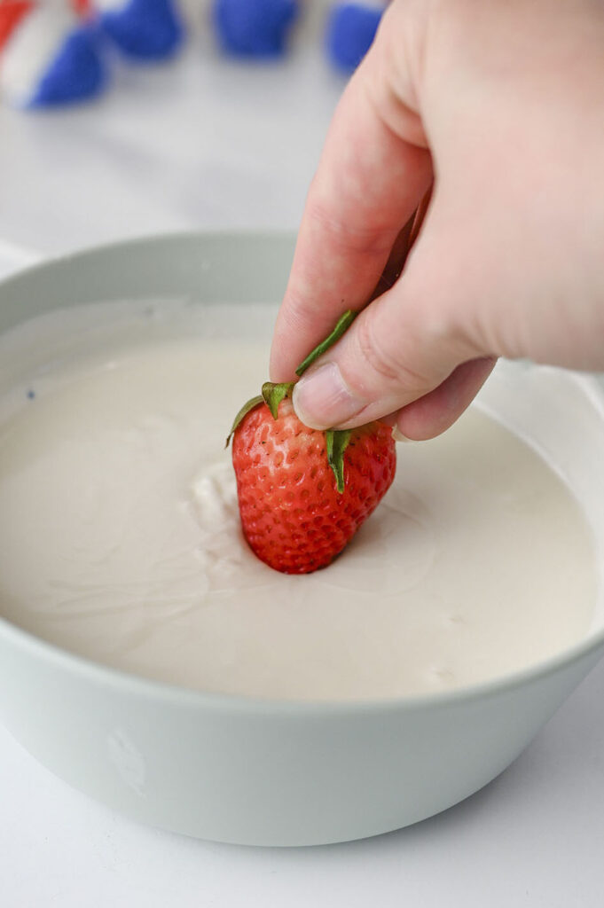 A hand dipping a fresh strawberry into a bowl of melted vanilla almond bark