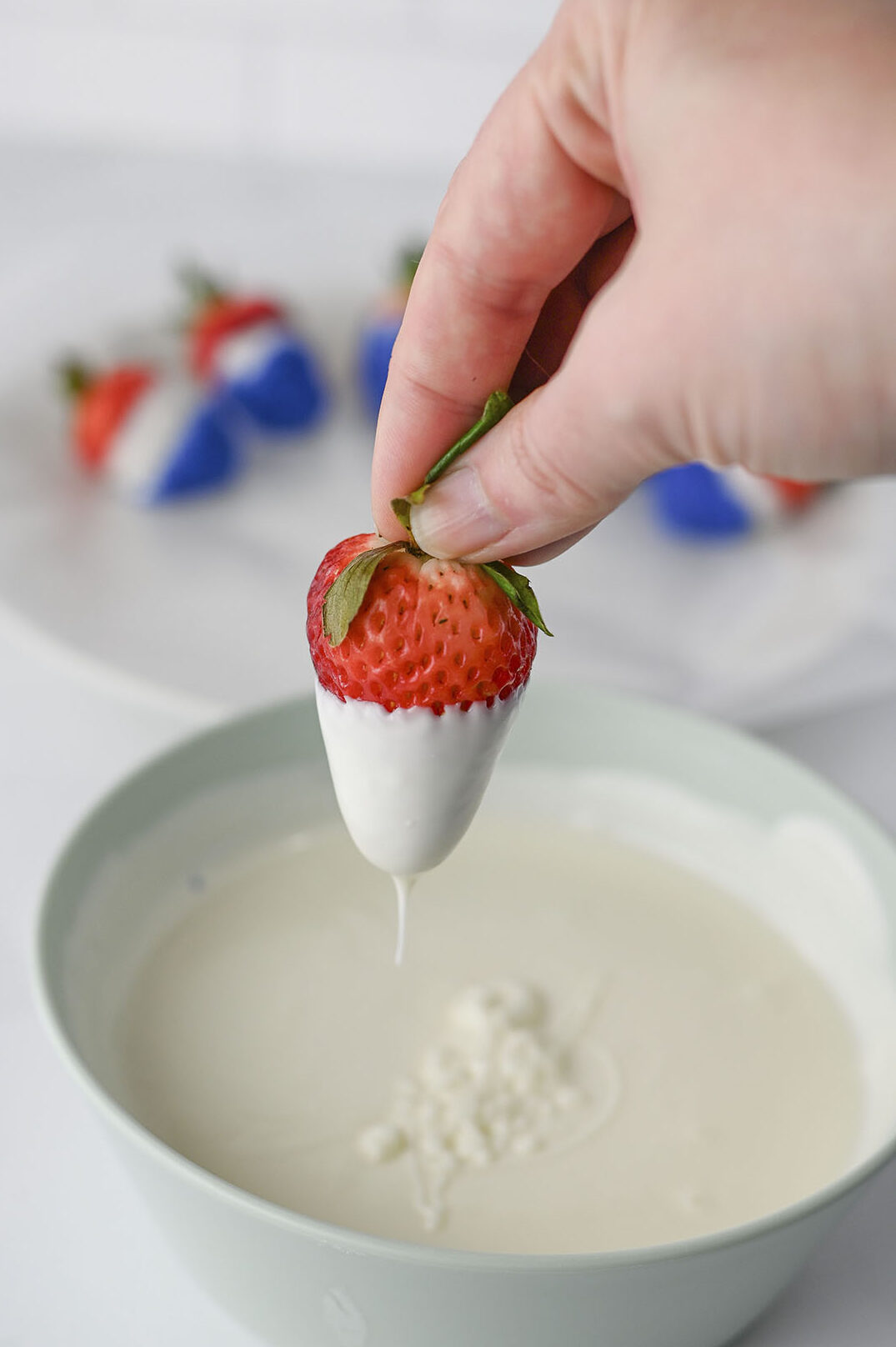 A hand holding a strawberry dipped in melted vanilla almond bark over a bowl.