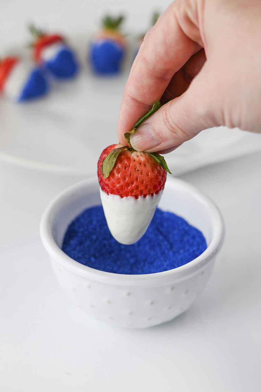 A hand holding a vanilla almond bark-dipped strawberry over a small bowl of blue sugar.